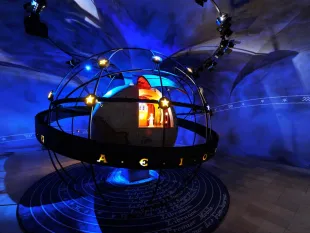 Ausstellungsraum: Eine leuchtende Kugel mit Sternen und einer Figur im Inneren, umgeben von einem Gestell und einem Boden mit astronomischen Beschriftungen.