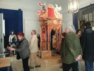 Ausstellungsraum: Prächtiger roter und goldener Altar mit Statue, umgeben von Besuchern und einem historisch gekleideten Wärter