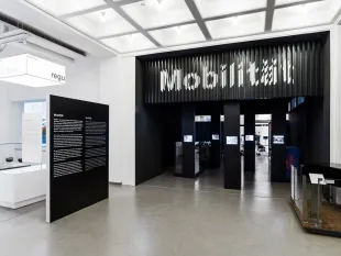 Ausstellungsraum: Moderner Raum mit der Aufschrift "Mobilität" über einem dunklen Eingang, umgeben von Informationspaneelen und Ausstellungsstücken.