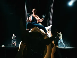 Szene: Schauspieler schwebt über der Bühne auf einer segmentierte Bullen-Skulptur, gehalten von Seilen. Zwei Darsteller um ihn herum