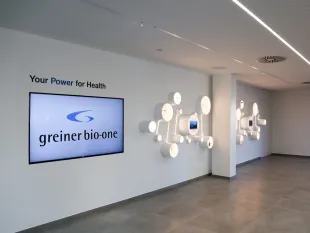 Ausstellungsraum: Ein Bildschirm mit "greiner bio-one" Logo, Text "Your Power for Health", und eine Wand mit beleuchteten, runden Elementen