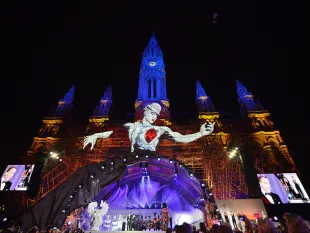 Wiener Rathaus mit beleuchteten Türmen, riesige Figur mit rotem Herzen, Bühne mit Performern, Publikum, Bildschirme, nächtliche Atmosphäre