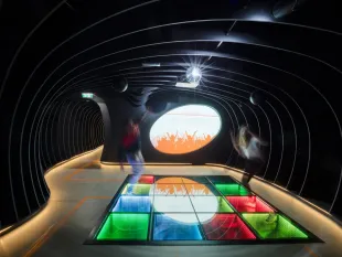 Ausstellungsraum: Tunnel mit gewölbter Decke, beleuchteter Boden mit bunten Lichtern, eine Projektion von tanzenden Silhouetten und Personen in Bewegung