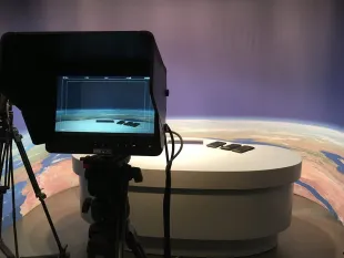 Studio mit einer großen LED-Wand im Hintergrund, die eine Erdkugel zeigt, einem höhenverstellbaren Pult mit Touchscreens und einer Kamera im Vordergrund.