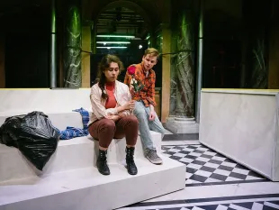 Zwei Personen auf weißen Treppen in dunklem Theaterraum, einer hält eine rote Rose, daneben Müllsack und Kleidungsstück.