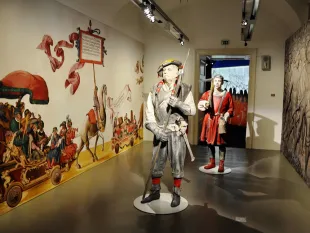 Ausstellungsraum: zwei Figuren in historischen Kostümen, ein Soldat mit Speer und ein rot gekleideter Mann, vor einer Wand mit farbenfroher Darstellung einer Prozession mit Pferden und Musikern