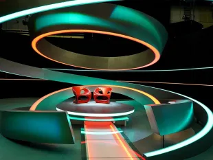 Bühne mit zwei roten Stühlen auf einer kreisrunden Plattform, umgeben von neonbeleuchteten, geschwungenen Elementen in Grün und Orange