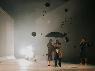 Zwei Darsteller unter schwarzem Regenschirm, umgeben von Nebel und schwarzen Luftballons