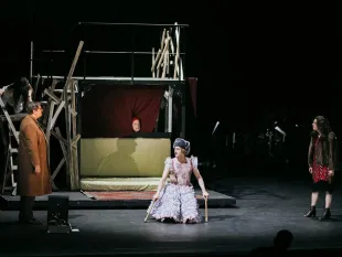 Schauspieler in historischem Kostüm mit Krücken auf Plattform, umgeben von Holzkonstruktionen und weiteren Figuren