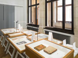 Ausstellungsraum mit Tischen, die historische Dokumente und Bücher in Vitrinen zeigen, vor großen Fenstern