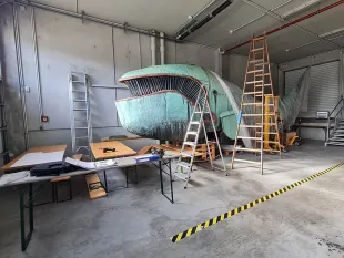 Grünes Wal-Modell (Praterwal „Poldi“) steht in einer Werkstatt, umgeben von Leitern und Werkzeugen. 