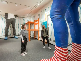 Ausstellungsraum mit zwei Frauen vor überlebensgroßen Figuren, die bis zur Decke reichen und nur teilweise sichtbar sind