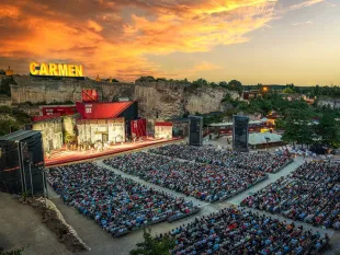 Open-Air-Bühne im Steinbruch mit einem großen „CARMEN“-Schriftzug an der Felskante, einer Filmset-Kulisse und Publikum bei Sonnenuntergang.