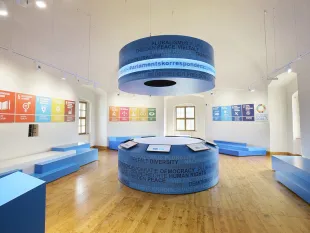 Ausstellungsraum mit einem zentralen, beleuchteten Rundtisch, einem hängenden Objekt darüber, umgeben von blauen Sitzflächen und Wänden mit thematischen Plakaten.