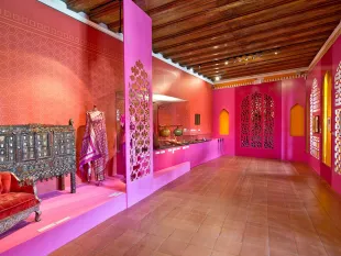Ausstellungsraum mit pinken Wänden und orientalischen Dekorationen, darunter ein aufwendig geschnitztes Möbelstück, ein farbenfrohes Kleid auf Ständer und verschiedene Kunstgegenstände in Vitrinen