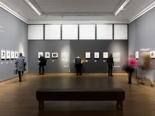 Ausstellungsraum: einige kleine Bilder an grauen Wänden, einige Besucher im Raum