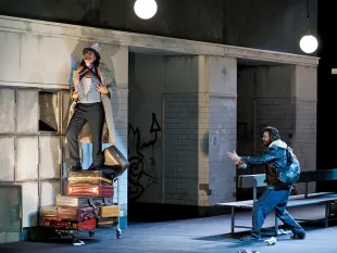 Szene: Mann in Mantel und Hut auf Kofferstapel, ein anderer in Lederjacke gestikuliert vor grauer Graffiti-Wand