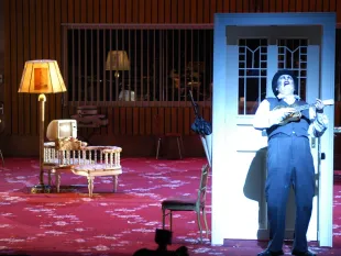 Szene: Mann steht mit Ukulele vor freistehnder Tür, roter Teppich mit alten Sesseln und Lampe im Hintergrund