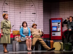 Szene: Drei Personen auf einer Bahnhofsbühne mit Uhr, Fahrkartenautomat und Gepäck; eine Frau steht, zwei sitzen, ein Mann mit Hut lehnt sich zurück, ein weiterer trinkt