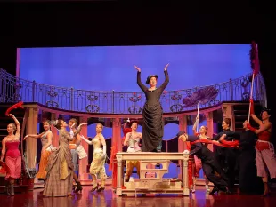 Szene: Frau steht auf einem Tisch mit erhobenen Armen, umgeben von tanzenden Darstellern  