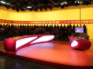 Studio mit roter Bühne, pinkfarbenem Pult und gelber Rückwand, beschriftet mit "Neues aus Was gibt es?", vor einem Publikum