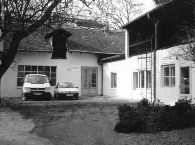 Werkstatt mit weißer Fassade, zwei geparkten Autos vor der Tür, einer Außenleiter und kahlen Bäumen, in Schwarz-Weiß fotografiert.