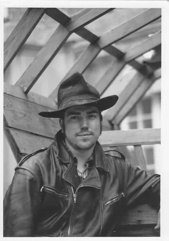 Mann in Lederjacke und Hut sitzt unter den Holzbalken eines Wagens in schwarz weiß fotografiert