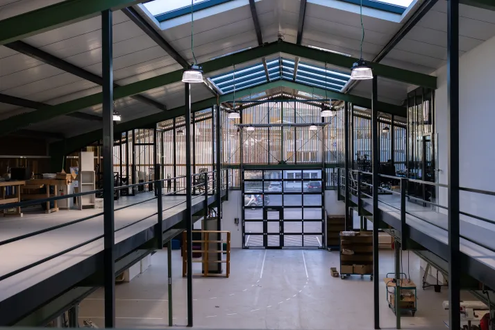 Werkstatt mit hoher Decke, Glasdach und Galerie, ausgestattet mit Arbeitsplätzen, Regalen und einem großen Glasportal, beleuchtet durch hängende Lampen.