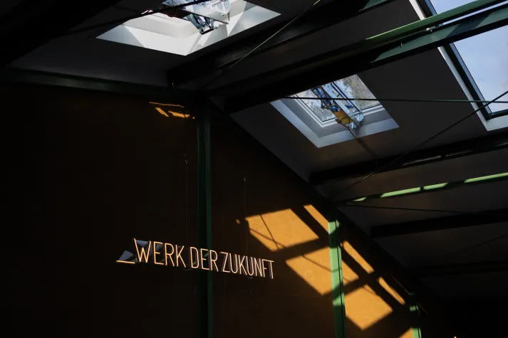 Eine Innenansicht einer Werkhalle mit hoher Decke, durch große Fenster beleuchtet und der leuchteter Aufschrift "Werk der Zukunft" an der Wand.