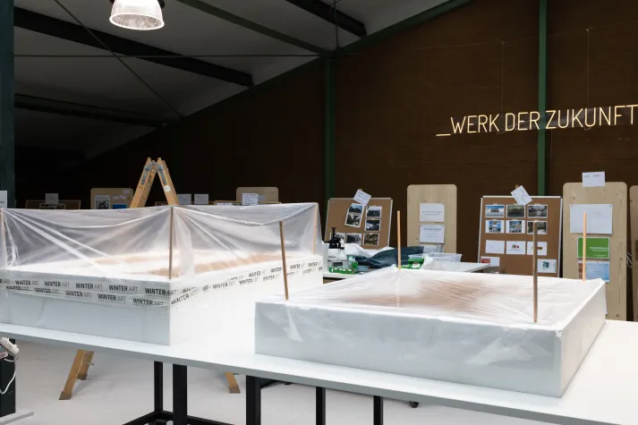 Zwei abgedeckte Styroblöcke auf einem Tisch in einer Werkhalle und der Aufschrift "Werk der Zukunft" an der Wand.