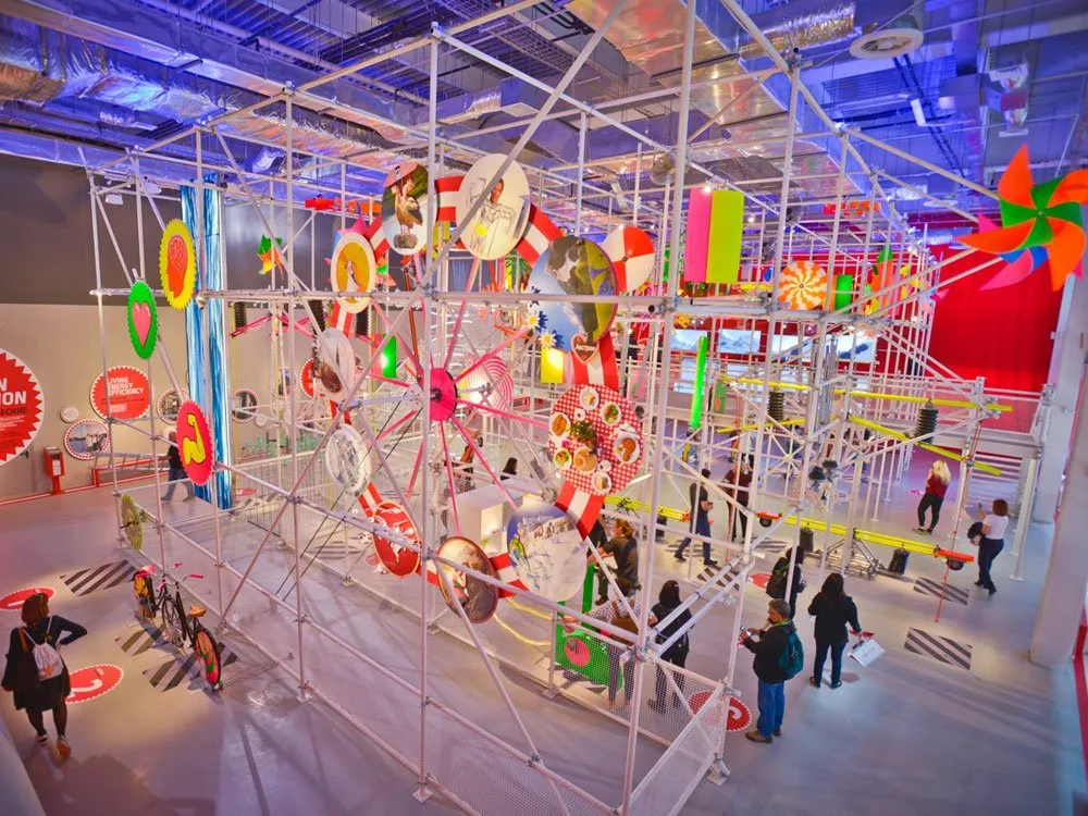 Farbenfrohe Installation mit Metallgerüst, bunten Ringen, Windrädern und Fahrrädern, umgeben von Besuchern