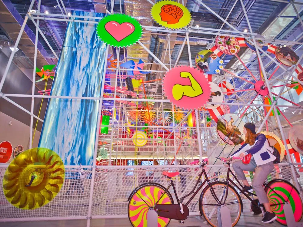 Bunte Installation mit Metallgerüst, Fahrrädern mit psychedelischen Rädern, großen dekorativen Symbolen (Herz, Muskel, Gehirn) und einem blauen Wasserfall-Element