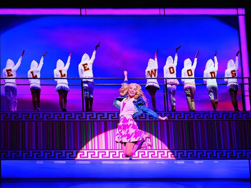 Szene: Frau mit blonden Haaren springt in pinkem Rock und Jeansjacke, acht Tänzerinnen in "ELLE WOODS"-Hoodies