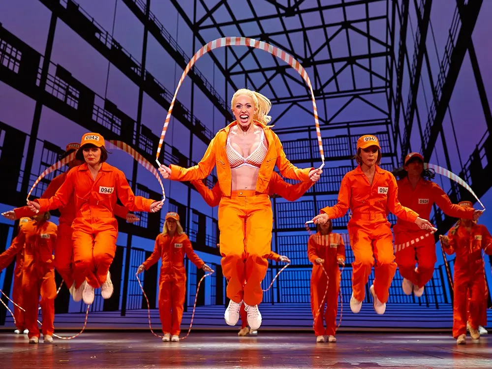 Szene: Darsteller in orangenen Overalls beim Seilspringen mit einem Absperrband