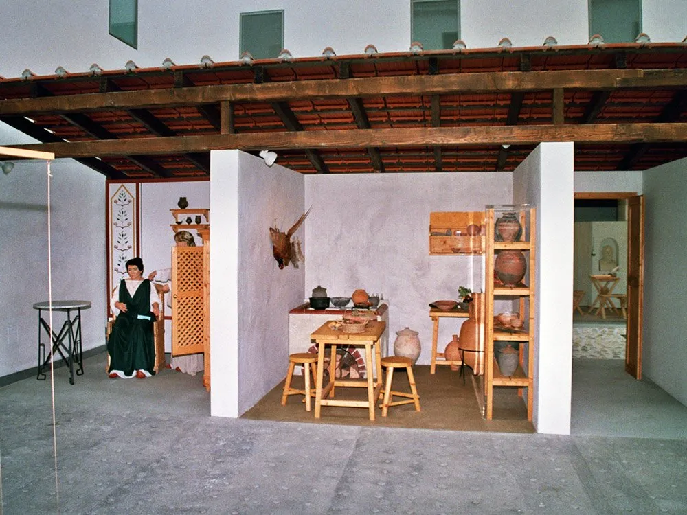 Nachbildung eines römischen Raumes mit sitzender Figur, Tisch, Töpfen und Regalen mit Keramiken unter Holzdach.