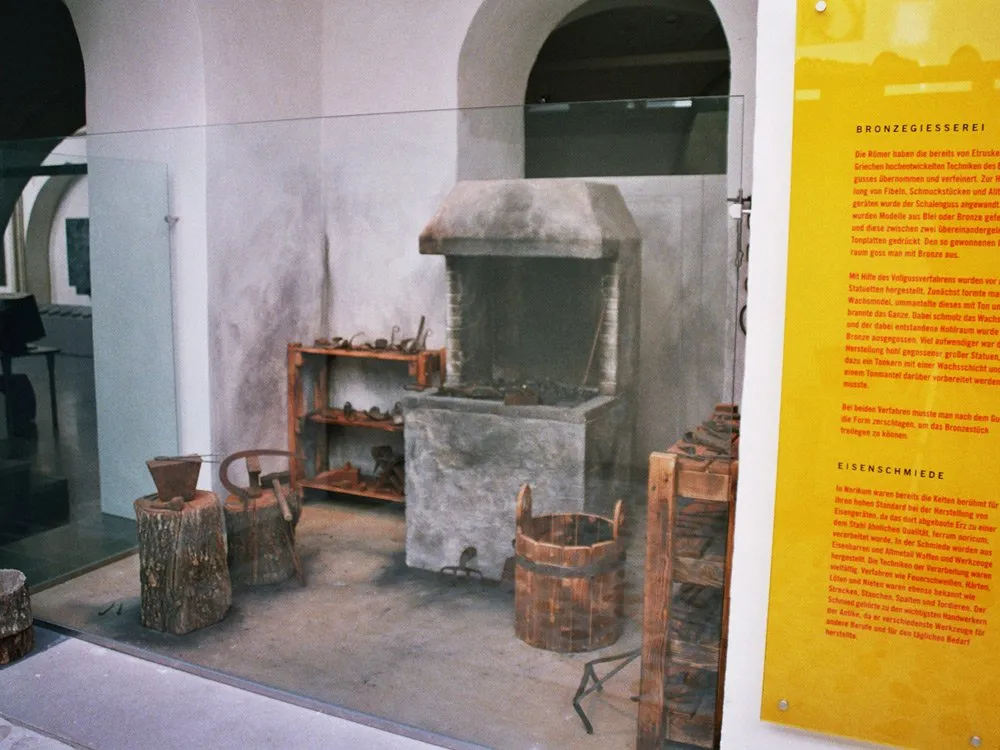 Nachbildung einer Bronze- und Eisenschmiede mit Ofen, Amboss, Werkzeugen und Holzfässern hinter Glas, begleitet von Informationsschildern