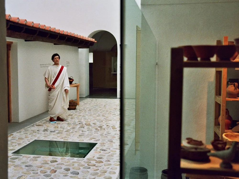 römische Figur in Toga auf einem gepflasterten Innenhof mit Glasboden, neben einem Regal mit Keramiken