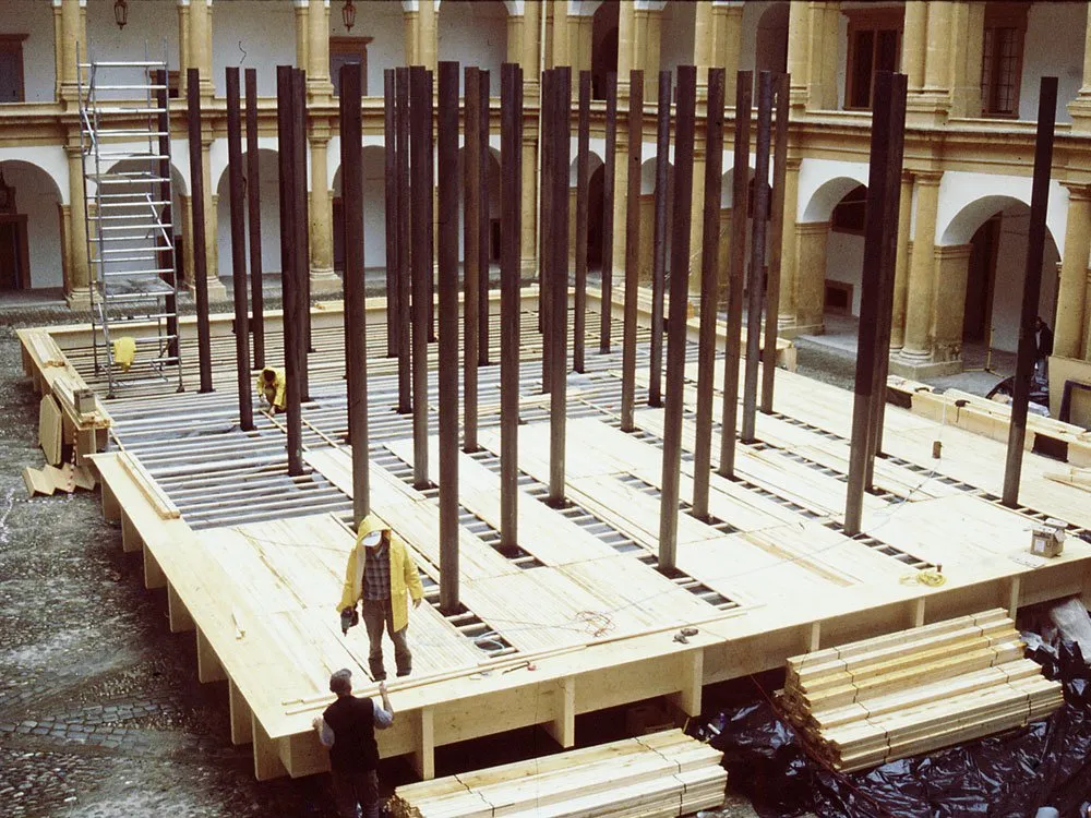 Ausstellungsraum: Holzpodest im Innenhof eines historischen Gebäudes mit Säulen, Arbeiter in gelben Regenmänteln beim Aufbau, Baugerüste und Materialien ringsum.