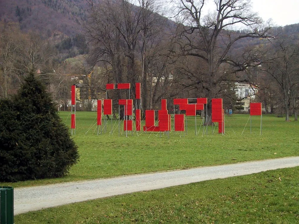 Ausstellungsraum: Grüne Wiese mit roten Platten auf Ständern, umgeben von kahlen Bäumen, Bergen und einem Kiesweg