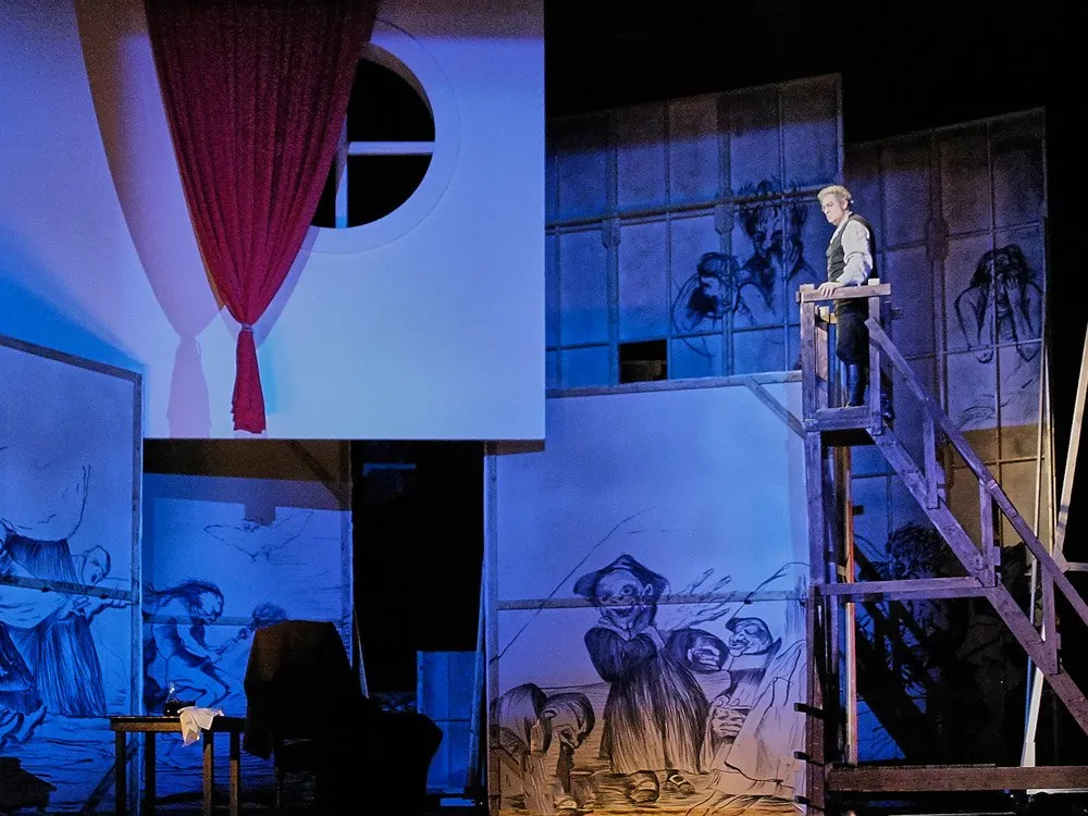 Szene: Schauspieler auf einer Leiter vor bemalten Wänden mit Figuren, einer roten Gardine und einem runden Fenster in blauem Licht