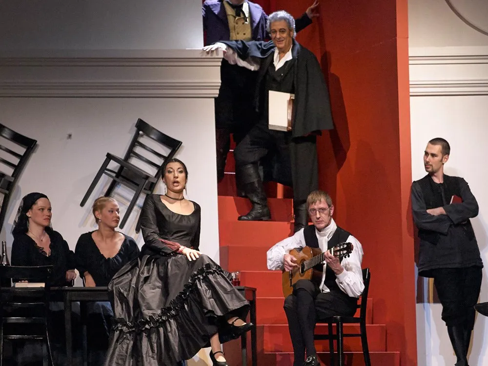 Schauspieler auf einer Treppe, einer Frau in schwarzem Kleid sitzend, einem Gitarristen und weiteren Personen, umgeben von umgekippten Stühlen und roter Kulisse