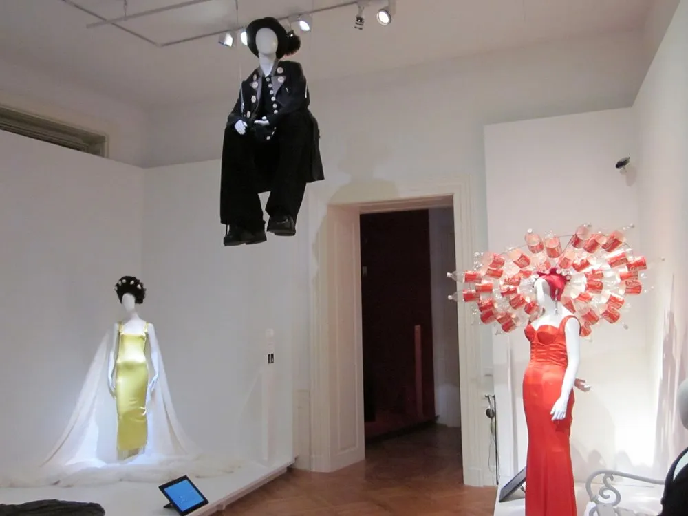 drei Schaufensterpuppen in fantasievollen Outfits – eine sitzt schwebend in schwarzer Kleidung mit Buttons, eine trägt ein gelbes Kleid mit weißem Umhang und Lockenwicklern, die dritte ein rotes Kleid mit einem auffälligen Kopfschmuck aus Coca-Cola-Flaschen.