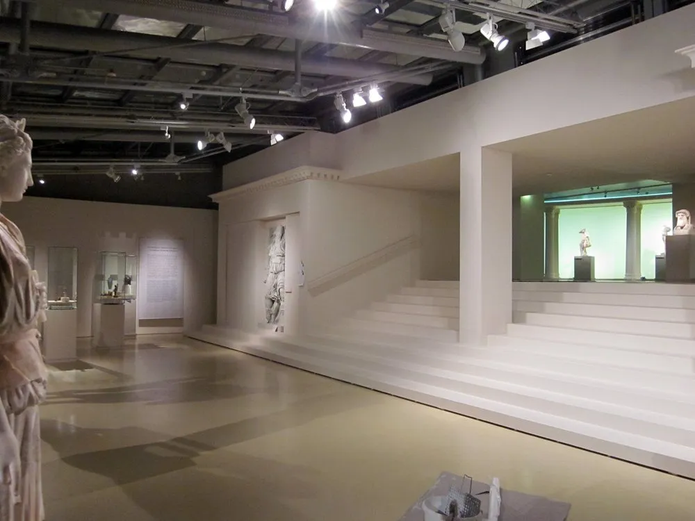Ausstellungsraum: Antike Skulpturen und Reliefs auf mehreren Ebenen, mit breiter Treppe und moderner Beleuchtung, eingerahmt von Vitrinen und klassizistischen Architekturelementen