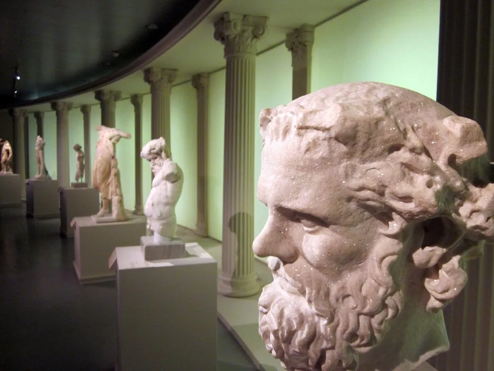 Ausstellungsraum: Eine Reihe antiker Marmorstatuen und -büsten, ausgestellt auf Podesten in einem Raum mit klassischen Säulen