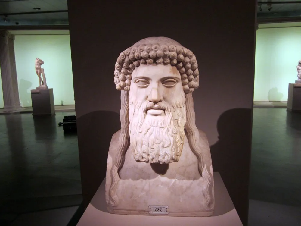 Ausstellungsraum: Marmorbüste mit Bart und lockigem Haar auf Podest, weitere Statuen und Säulen im Hintergrund.