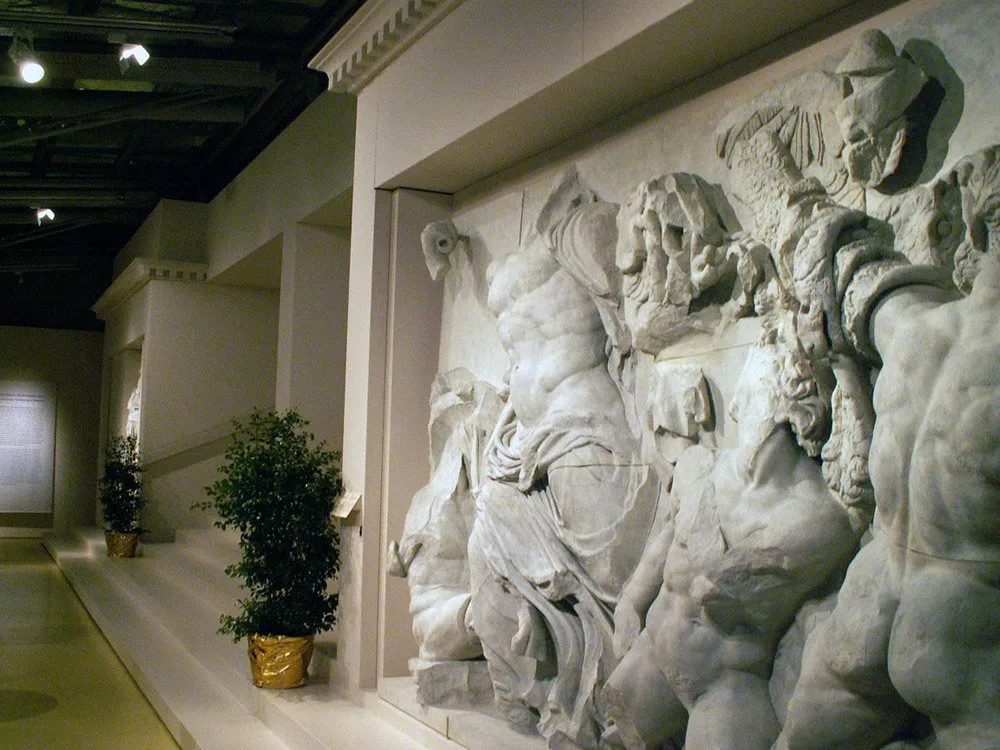 Ausstellungsraum: Marmorrelief mit Figuren an der Wand, Treppen, Säulen und goldene Topfpflanzen im Hintergrund