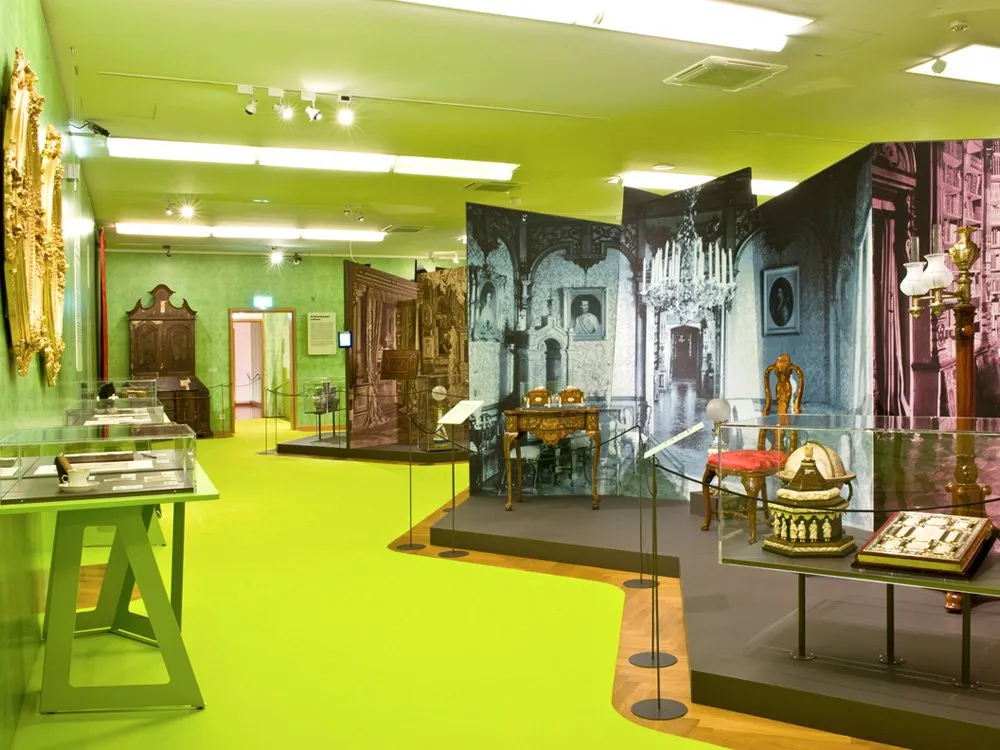 Ausstellungsraum: Grüne Wände und Boden, Vitrinen mit historischen Objekten und Möbeln, Wandbilder mit antiken Szenen