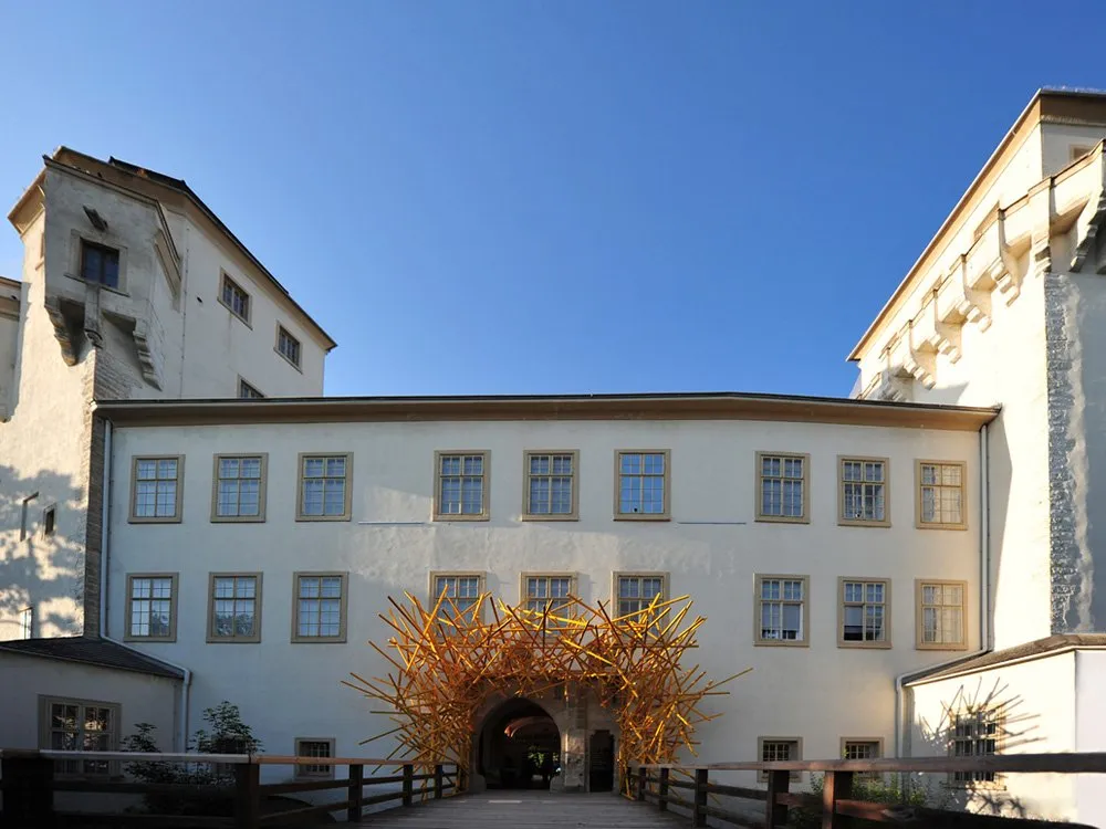 Ausstellungsraum: Weißes Gebäude mit gelber Skulptur über dem Eingang, umgeben von Fenstern mit grauen Rahmen, Holzbrücke davor, blauer Himmel im Hintergrund.