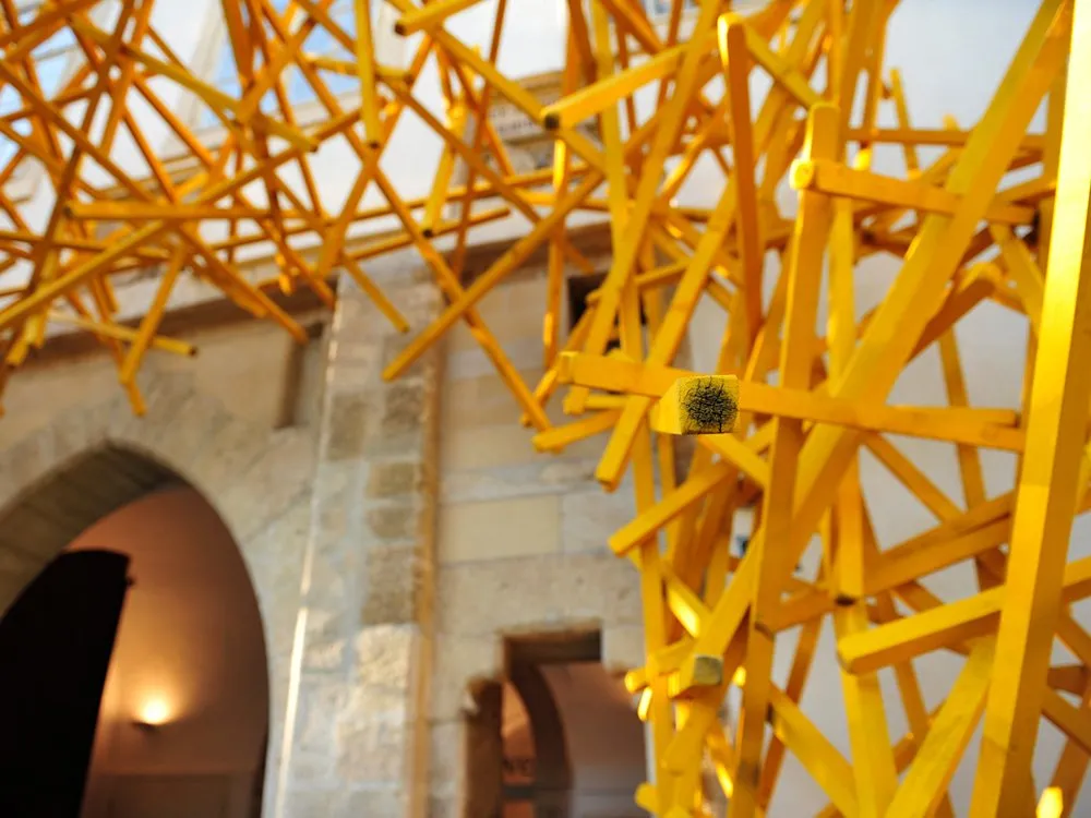 Ausstellungsraum: Gelbe Skulptur aus Holzstäben über einem steinernen Torbogen, beleuchteter Innenraum im Hintergrund.