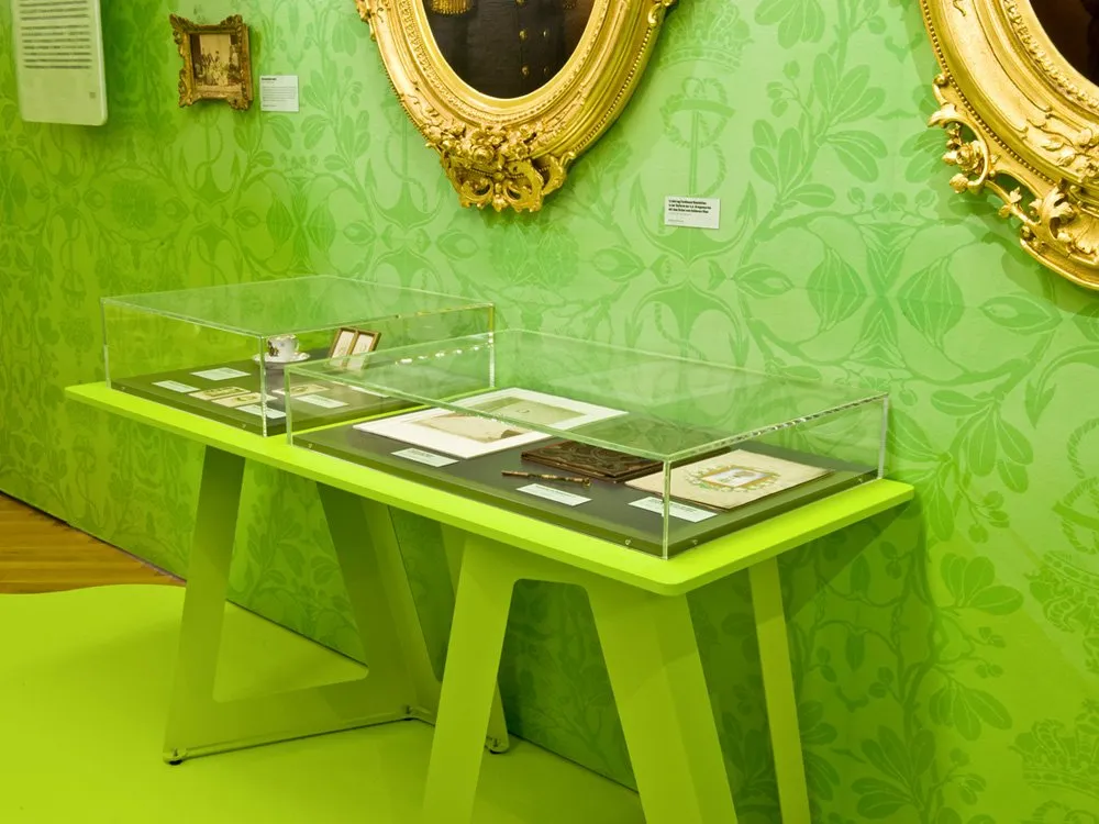 Ausstellungsraum: Grün gestalteter Raum mit floraler Tapete, goldgerahmten Porträts und Vitrine mit historischen Artefakten